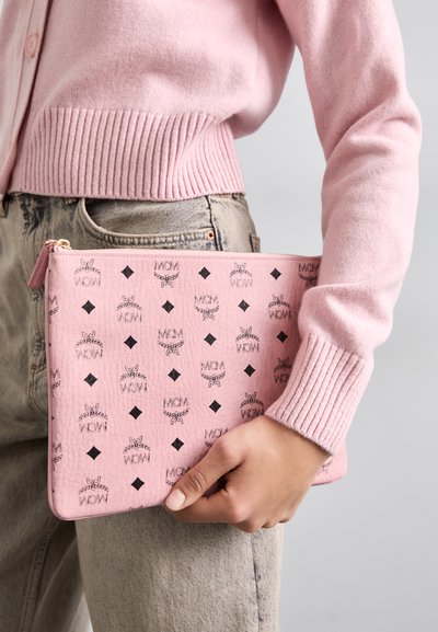 Pungă din piele roz cu logo negru și model în diamant. Punga are un închizător tip fermoar și este ținută de o persoană îmbrăcată cu un cardigan roz.
