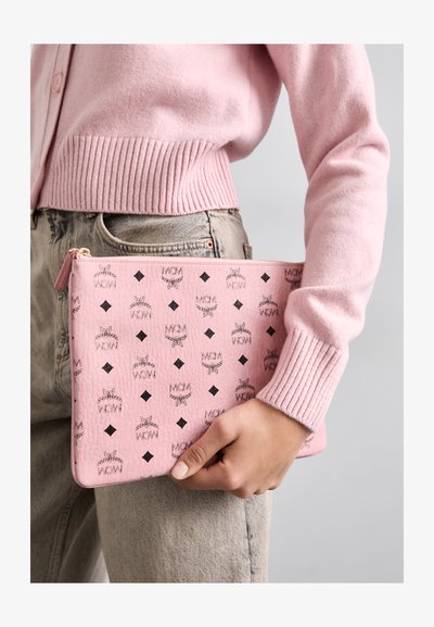 Pungă din piele roz cu logo negru și model în diamant. Punga are un închizător tip fermoar și este ținută de o persoană îmbrăcată cu un cardigan roz.