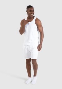 Weißes Tanktop und Shorts-Set, aus leichtem Stoff. Das Model steht lächelnd da, trägt weiße Knöchelsocken, vor einem neutralen Hintergrund.
