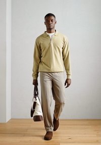 Beige cipzáros pulóver, világos bézs nadrág, barna csizma, egy krémszínű és barna táskával a kezében. Sima anyag, laza szabás, minimalista design.