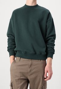 Man in een donkergroen sweatshirt met ronde hals en lichtbruine cargobroek, met één hand in zijn zak en de andere ontspannen naast zijn lichaam.