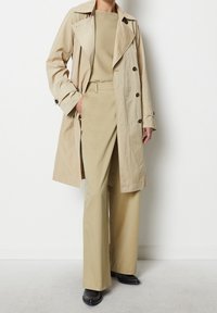 Trench coat beige con design doppiopetto, top beige abbinato e pantaloni a gamba larga. Tessuto morbido e dettagli con bottoni. Scarpe nere.
