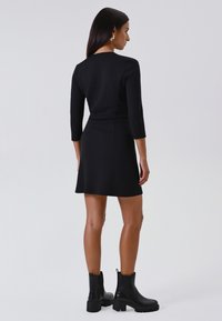 Schwarzes, tailliertes Kleid mit Dreiviertelärmeln und ausgefranstem Saum, kombiniert mit schwarzen Ankle Boots mit dicken Sohlen. Glattes Material und klare Linien.