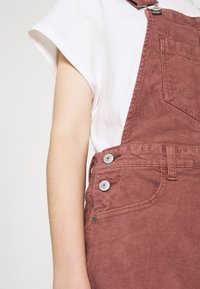 Salopette en velours côtelé rouge rouille avec détails métalliques argentés. Comprend une poche avant et des bretelles réglables. Associée à un t-shirt blanc uni.