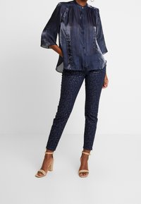 Blouse en satin bleu marine à encolure ronde avec détails plissés, associée à un pantalon ajusté à imprimé léopard bleu foncé et des sandales à talons blocs beige.