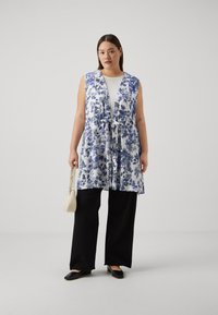 Fehér virágmintás mellény kék mintákkal, derékon megkötő rész. Fekete, bő szárú nadrággal és fekete slip-on cipővel párosítva. Minimalista kézitáska.