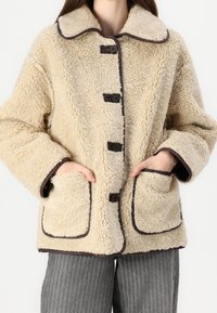 Beige tekoturkistakki, jossa suuri kaulus, viisi mustaa nappia ja ruskea reunus. Takin etuosassa on kaksi taskua ja rento istuvuus.