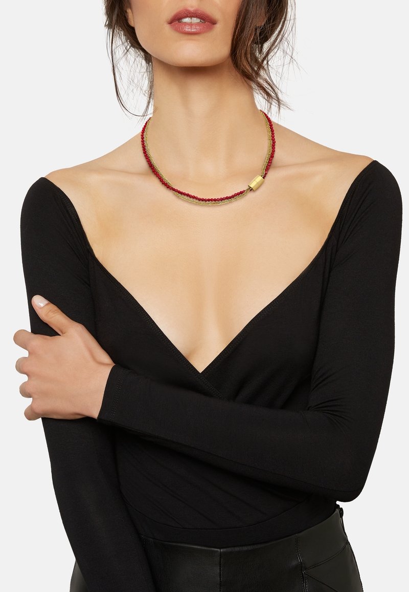 Femme portant un haut noir à décolleté plongeant en V et un collier à double rang avec des perles rouges et une chaîne dorée, bras croisés sur la poitrine.