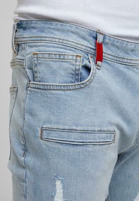 Világoskék farmernadrág mintázott befejezéssel, két hátsó zsebbel és egy piros címkével az elülső zsebben. A varrás kontrasztos cérnával készült.