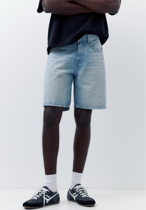 Person trägt hellblaue Jeansshorts, schwarz-weiße Sneakers, weiße Socken und ein schwarzes Shirt mit verschränkten Armen.