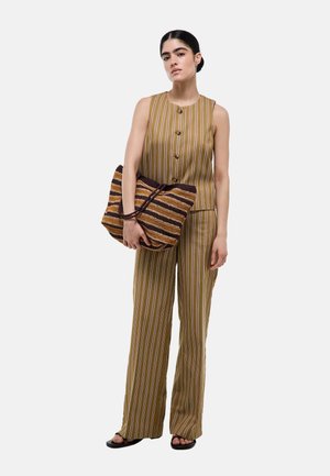 Femme portant un haut marron rayé sans manches boutonné et un pantalon large assorti, tenant un grand sac tissé rayé et portant des sandales.