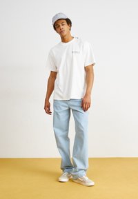 White cotton t-shirt with a small graphic, light blue denim jeans, and beige sneakers. The model wears a blue and white striped cap.