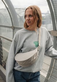 Giovane donna in felpa grigia con testo verde e borsa a tracolla beige si trova in un corridoio chiuso, guardando di lato all'aperto.