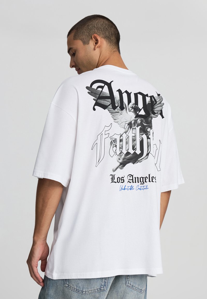 T-shirt bianco in cotone con grafica testuale "Fede nella Rabbia" e un design di un angelo con ali sul retro. Vestibilità ampia con maniche corte.