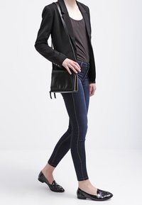Blazer noir sur un haut gris, associé à un jean skinny foncé et des mocassins en cuir verni noir. Le modèle porte un sac bandoulière noir.