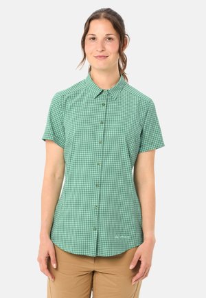 Frau, die ein grünes, kariertes Kurzarm-Button-up-Hemd und beige Hosen trägt, steht vor einem schlichten weißen Hintergrund.