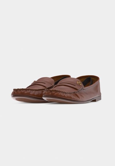 Mocassins en cuir marron avec des bouts arrondis cousus et un petit emblème doré sur le dessus, placés de côté l'un à côté de l'autre sur un fond blanc uni.
