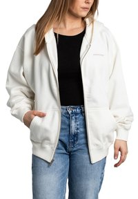 Bílé zipové mikina s kapucí, předními kapsami a žebrovanými manžetami. Nosená přes černý top, doplněná o vysoké modré džíny.