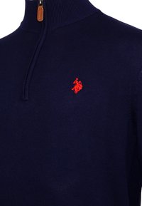 Marineblaues Quarter-Zip-Sweatshirt mit einem roten, gestickten Polo-Spieler-Logo auf der linken Brust und einem Reißverschlussanhänger aus braunem Leder.