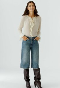 Cremfarbene Chiffonbluse mit Rüschendetails, kombiniert mit hochgeschnittenen, hellblauen Schlagjeans und kniehohen, braunen Stiefeln.