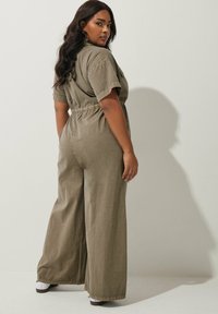 Kakifärgad jumpsuit med korta ärmar, figursydd midja och vida byxor. Materialet verkar lätt och har en strukturerad yta.