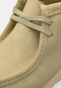 Beige suede shoe with a rounded toe, featuring decorative stitching and thin, crisscrossed laces. Smooth, textured surface detail.
