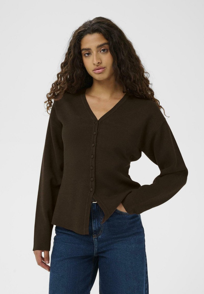 Brūns kardigāns ar V veida kakla izgriezumu, pogu priekšpusi un garām piedurknēm. Mīksta tekstūra un sānu šķēlumi; savienots ar ziliem augstiem džinsiem.