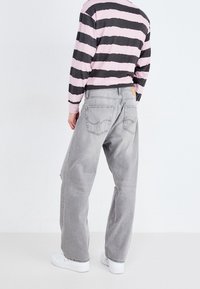 Jeans grigi oversized con una vestibilità larga, dotati di tasche posteriori, orli sfilacciati, abbinati a sneakers bianche e a una maglietta a righe rosa e nera.