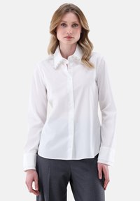 Camicia bianca con colletto, impreziosita da dettagli simili a perle, maniche lunghe e una consistenza liscia. Indossata con pantaloni grigi.