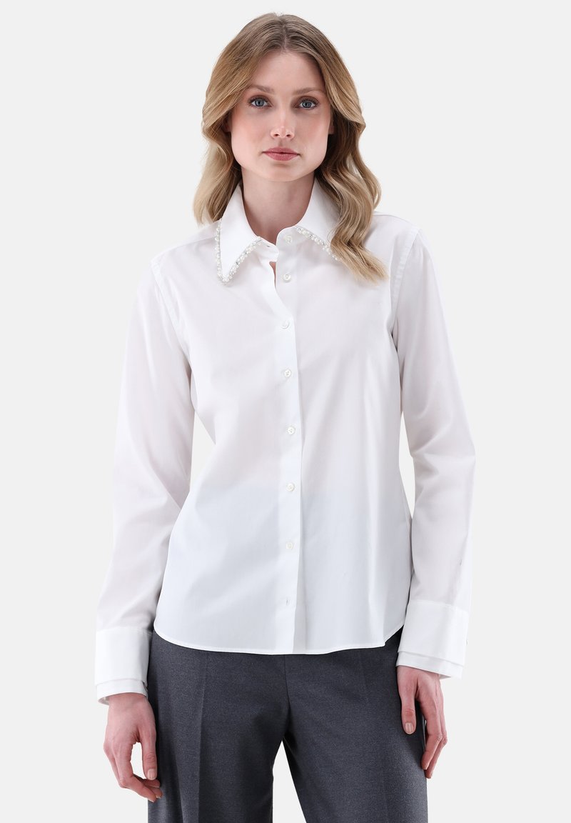 Camicia bianca con colletto, impreziosita da dettagli simili a perle, maniche lunghe e una consistenza liscia. Indossata con pantaloni grigi.
