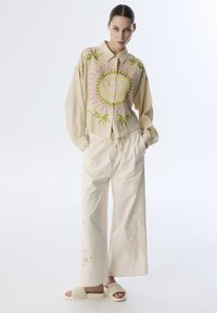 Camisa corta beige con bordados en verde y rosa, combinada con pantalones de pierna ancha crema que presentan sutiles patrones de peces, y sandalias trenzadas crema.
