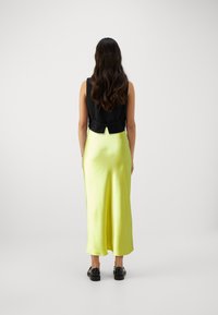 Haut sans manches noir avec détail de nœud associé à une jupe en satin jaune vif longue jusqu'au sol. Le modèle porte des chaussures noires à lacets.
