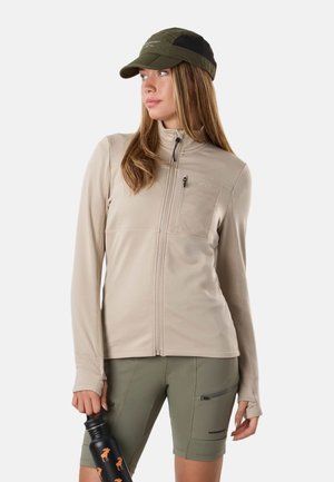 Kobieta ubrana w beżową kurtkę na zamek, oliwkowe szorty i zieloną czapkę, trzyma czarną butelkę na wodę z pomarańczowymi sylwetkami zwierząt.