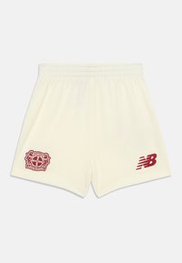 Calções de cor creme feitos de tecido suave, apresentando um brasão de clube vermelho na perna esquerda e um logótipo vermelho da New Balance na perna direita.
