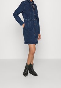 Kobieta w zapinanej ciemnoniebieskiej sukience dżinsowej z długimi rękawami i zielonych botkach z kroko wzorem do kostek, stojąca na jednolitym tle.