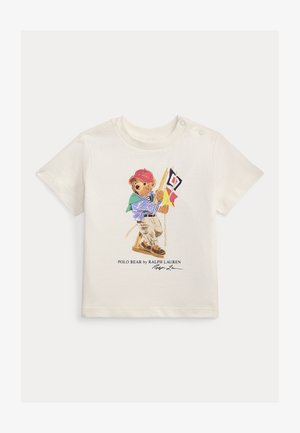 Baltas kūdikio marškinėlių palaidinė su meškučiu, dėvinčiu raudoną kepuraitę, mėlynai dryžuotą marškinius, smėlio spalvos kelnes, laikantį jūrinius signalinius vėliavėlius sūpynėse.