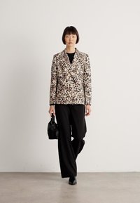 Vrouw in een blazer met luipaardprint, zwarte wijde broek en zwarte schoenen, met een kleine zwarte handtas, loopt naar voren.