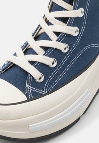 Närbild av en blå canvas-sneaker med vita snörningar, vit gummitå, svart yttersula och detaljer i vit sömnad.