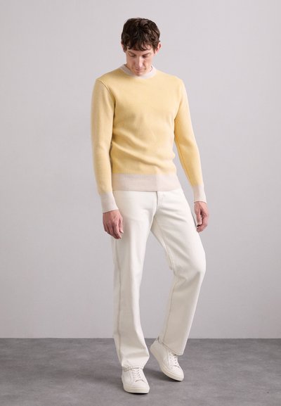Gelbes, geripptes Strickoberteil mit einem hellbeigen Saum, kombiniert mit weißen Jeans und weißen Sneakers. Modell posiert vor einem neutralen Hintergrund.