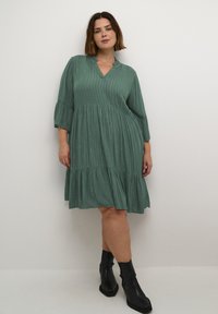 Vestido verde hasta la rodilla con rayas verticales, mangas acampanadas y un dobladillo con capas. Hecho de una tela suave con un acabado ligeramente texturizado.