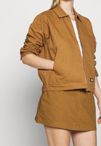 Femme portant une veste marron zippée avec les manches retroussées et une jupe marron assortie, la main posée de manière décontractée dans la poche de la veste.