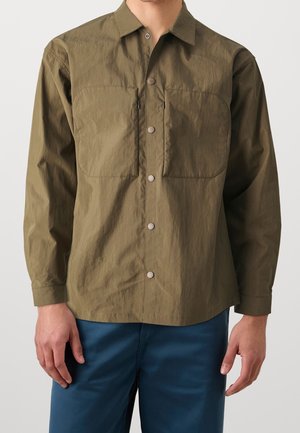 Mies, joka pukeutunut oliivinvihreään, pitkähihainen napilliseen paitaan, jossa on kaksi rintataskua, ja sinisiin housuihin yksinkertaista taustaa vasten.