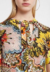 Blommönstrad topp med nyckelhålsutskärning. Färgerna inkluderar gult, rosa och grönt. Har texturerad tyg och korta  ärmar.