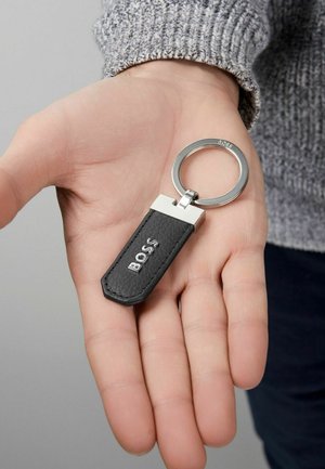 Black leather keychain with a silver metal ring, featuring a hexagonal shape with embossed logo detail. Handheld against a light background.