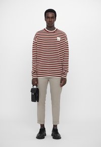 Gestreifter brauner und weißer Pullover mit Rundhalsausschnitt, kombiniert mit hellbeigen Hosen. Hält eine kleine schwarze Tasche. Schwarze Schuhe vervollständigen das Outfit.