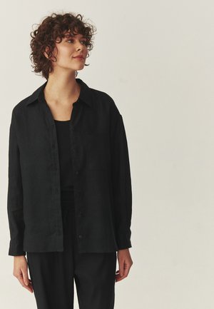 Młoda kobieta z kręconymi włosami, ubrana w dużą czarną koszulę na guziki i czarne spodnie, lekko patrząca na bok.