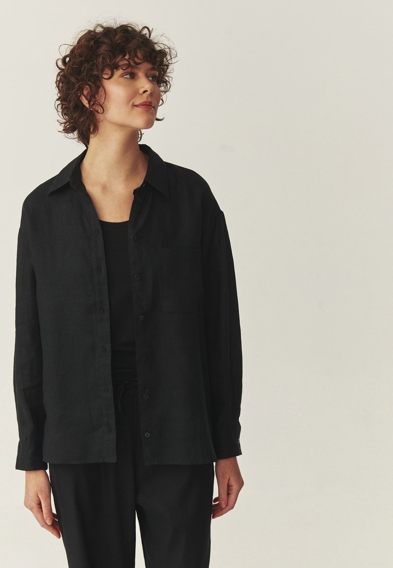 Jeune femme aux cheveux bouclés portant une chemise noire oversize boutonnée et un pantalon noir, regardant légèrement sur le côté.