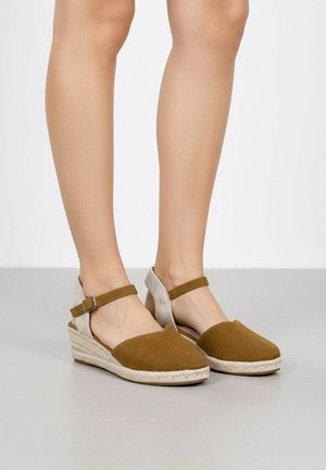 Jambes portant des sandales espadrilles compensées en daim marron avec des brides à la cheville et un détail tissé beige au talon sur un sol gris.