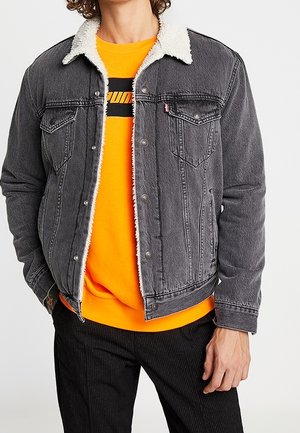 Osoba ubrana w szarą kurtkę dżinsową z białym kołnierzem sherpa na jasno-pomarańczowej bluzie z czarnym napisem oraz czarne spodnie.