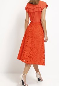 Orange blus med frillad rygg, parat med en djärv orange blyertskjol med blommig spetsutskärningar, kompletterat med vita högklackade skor.